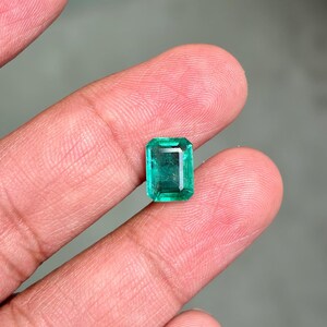 May include: A rectangular emerald gemstone with a vibrant green hue. The gem is held between fingers, showcasing its clarity and cut. The emerald's facets reflect light, highlighting its brilliance. This gemstone is a beautiful example of natural emeralds.