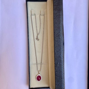 May include: A silver necklace with a round, red pendant surrounded by small, clear stones, displayed in an open black jewelry box with a cream-colored interior. The necklace chain is delicate and hangs gracefully.
