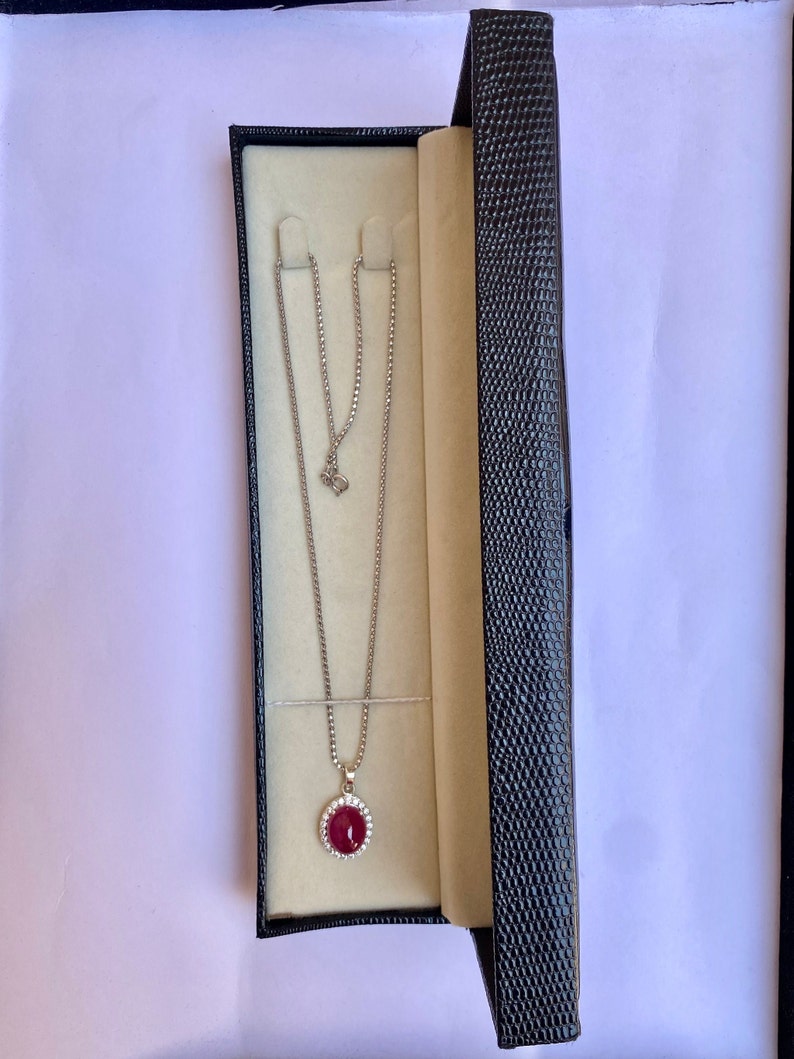 May include: A silver necklace with a round, red pendant surrounded by small, clear stones, displayed in an open black jewelry box with a cream-colored interior. The necklace chain is delicate and hangs gracefully.
