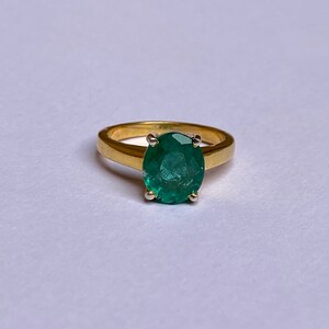 May include: A gold ring with a large, oval-shaped emerald gemstone set in the center. The ring has a simple band and the emerald is a vibrant green color.