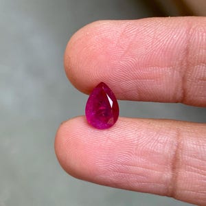 Könnte beinhalten: Ein facettierter, birnenförmiger Rubin-Edelstein. Der Edelstein hat eine tiefe, leuchtende rote Farbe, wobei mehrere Facetten das Licht einfangen. Der Rubin wird zwischen zwei Fingern gehalten, um seine Größe und Form zu zeigen. Ideal für die Schmuckherstellung.