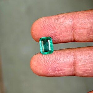 May include: A vibrant, emerald-cut green gemstone. The rectangular gem is held between two fingers, showcasing its clarity and color. The stone's facets reflect light, highlighting its brilliance. The background is a soft, neutral tone.