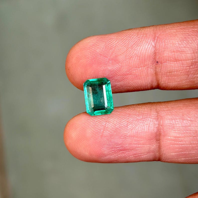 May include: A vibrant, emerald-cut green gemstone. The rectangular gem is held between two fingers, showcasing its clarity and color. The stone's facets reflect light, highlighting its brilliance. The background is a soft, neutral tone.