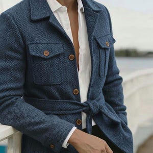 Puede incluir: Una chaqueta azul marino con diseño de espiga y cinturón a juego atado a la cintura. La chaqueta tiene dos bolsillos delanteros y botones de madera. Debajo se lleva una camisa blanca con cuello.