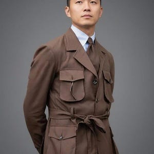 May include: A brown safari jacket with multiple pockets and a matching tie. The jacket has a belted waist and is worn over a striped shirt. The outfit is a classic style, suitable for a variety of occasions.