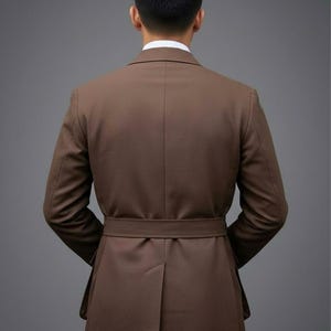 May include: A brown suit jacket with a matching belt, viewed from the back. The jacket has a tailored fit, with a single vent in the back. The person is wearing a white collared shirt underneath the jacket.