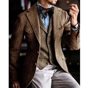 Puede incluir: Un traje de tres piezas de tweed marrón, que incluye una chaqueta con dos bolsillos delanteros y botones marrones. El chaleco tiene un diseño abotonado, y el traje se combina con una camisa azul claro y una pajarita marrón oscuro. Los pantalones son de color gris claro.