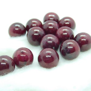 May include: A group of 13 deep red round beads, some with a slight shine, on a white surface.