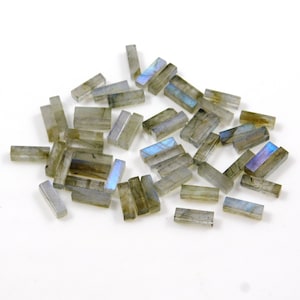 以下が含まれることがあります： 長方形の虹色のグレーのラブラドライトジェムストーンビーズのコレクション。ビーズは白い表面に散らばっています。