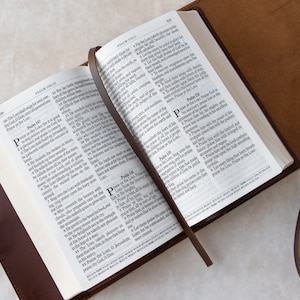 May include: A brown leather-bound Bible with a bookmark, open to Psalms 149 and 150. The pages are white with black text. The Bible is lying on a white surface.