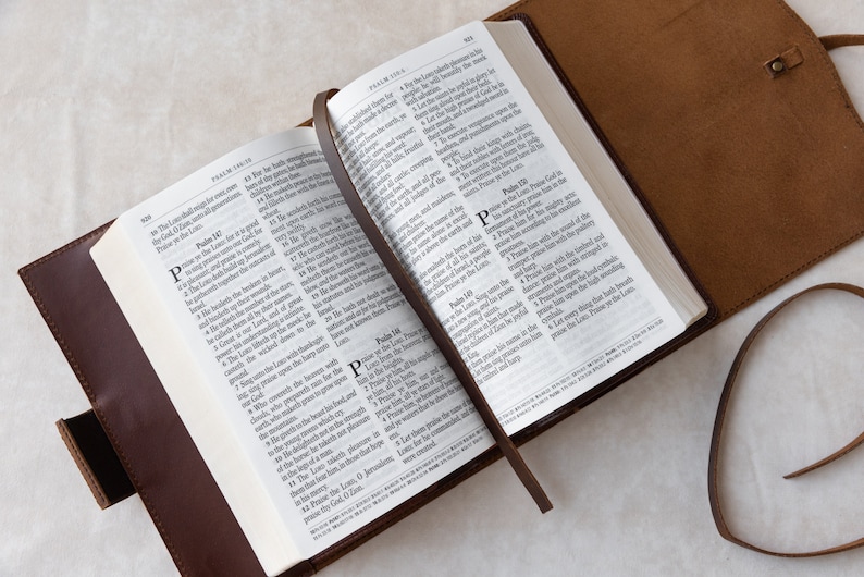 May include: A brown leather-bound Bible with a bookmark, open to Psalms 149 and 150. The pages are white with black text. The Bible is lying on a white surface.
