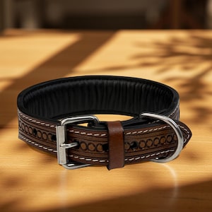 Op de afbeelding: Een bruine leren hondenhalsband met een zwarte gewatteerde binnenkant. De halsband heeft decoratieve ge&euml;tste patronen en witte stiksels. Het heeft een zilverkleurige metalen gesp en een bruine leren lus.