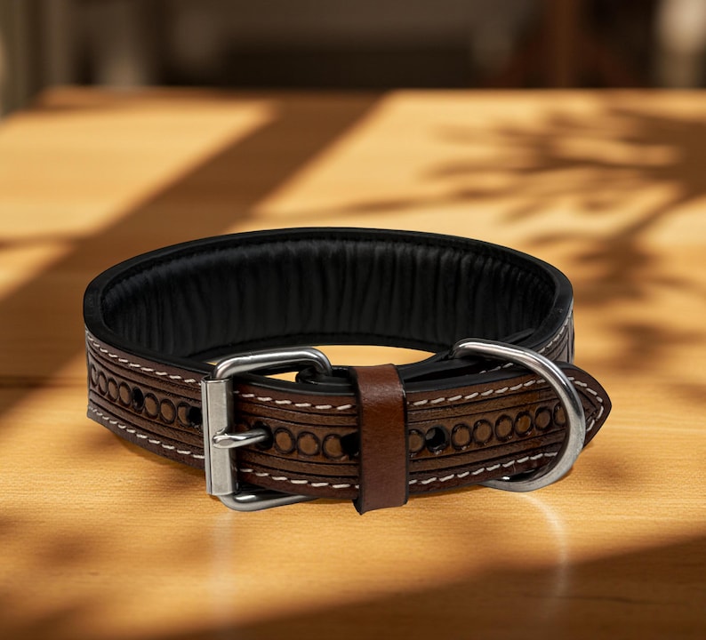 Op de afbeelding: Een bruine leren hondenhalsband met een zwarte gewatteerde binnenkant. De halsband heeft decoratieve ge&euml;tste patronen en witte stiksels. Het heeft een zilverkleurige metalen gesp en een bruine leren lus.