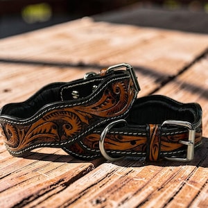 Peut inclure: Deux colliers de chien en cuir marron avec des motifs floraux complexes et des coutures blanches. Les colliers ont un dos noir et des boucles et anneaux en D en m&eacute;tal argent&eacute;. Ils sont pr&eacute;sent&eacute;s sur une surface en bois.