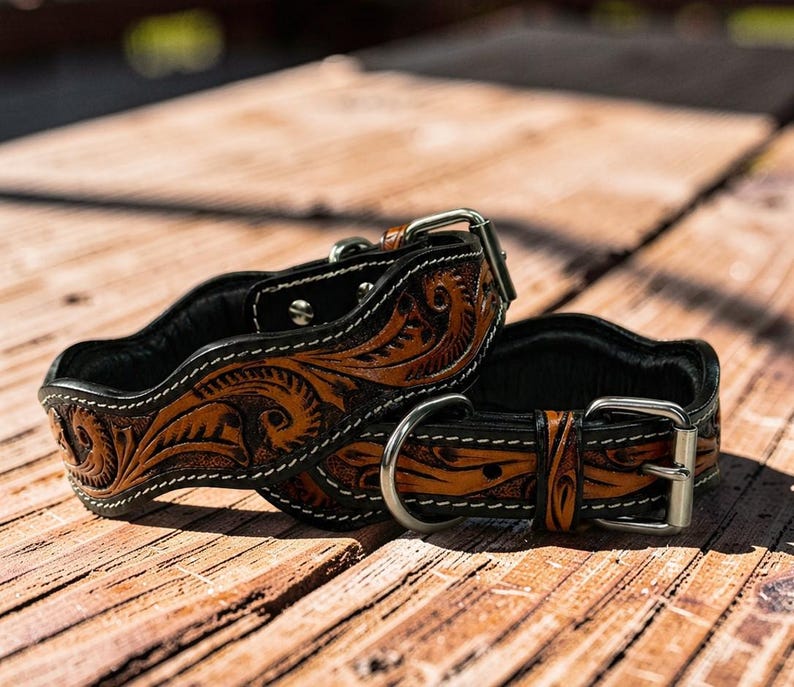 Peut inclure: Deux colliers de chien en cuir marron avec des motifs floraux complexes et des coutures blanches. Les colliers ont un dos noir et des boucles et anneaux en D en m&eacute;tal argent&eacute;. Ils sont pr&eacute;sent&eacute;s sur une surface en bois.
