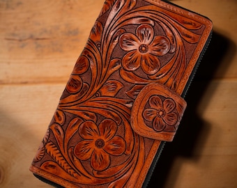 Puede incluir: Un monedero de cuero marrón con un diseño floral en relieve. El monedero tiene un cierre de cremallera y un cierre de botón a presión con un diseño de flor. La intrincada talla y el trabajo de herramientas crean una superficie detallada y texturizada.