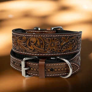 Op de afbeelding: Twee bruine leren hondenhalsbanden met bloemen- en bladeren tooling. De halsbanden hebben zilverkleurige gespen en witte stiksels. De bovenste halsband is bovenop de andere gestapeld. De achtergrond is een vage gradi&euml;nt van bruin en goud.