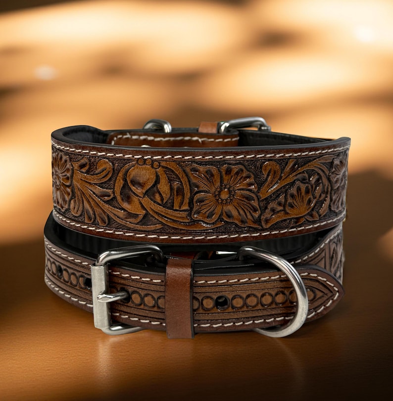 Op de afbeelding: Twee bruine leren hondenhalsbanden met bloemen- en bladeren tooling. De halsbanden hebben zilverkleurige gespen en witte stiksels. De bovenste halsband is bovenop de andere gestapeld. De achtergrond is een vage gradi&euml;nt van bruin en goud.