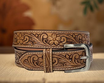 Op de afbeelding: Een gestapelde, lichtbruine leren riem met bloemen- en krulwerk. De riem heeft een zilverkleurige gesp en witte stiksels. Het ontwerp is een klassieke Western-stijl.