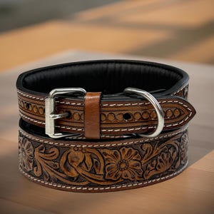 Op de afbeelding: Afbeelding van twee bruin lederen hondenhalsbanden, de ene bovenop de andere gestapeld. De bovenste halsband heeft een bruin lederen riem en zilveren hardware. De onderste halsband heeft een bloemreli&euml;f met witte stiksels.