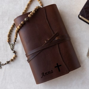 May include: A brown leather journal with a cross and the name "Anna" engraved on the front. A wooden rosary with a silver crucifix is lying on top of the journal.
