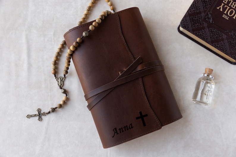 May include: A brown leather journal with a cross and the name "Anna" engraved on the front. A wooden rosary with a silver crucifix is lying on top of the journal.