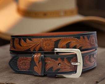 Op de afbeelding: Een bruine leren riem met ingewikkelde bloemmotieven en een zwarte rand. De riem heeft een zilverkleurige gesp en een klassiek western ontwerp. De gedetailleerde tooling toont traditioneel vakmanschap.