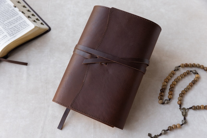May include: A brown leather-bound journal with a tied closure and a long brown bookmark. The journal is on a white surface with a wooden rosary.