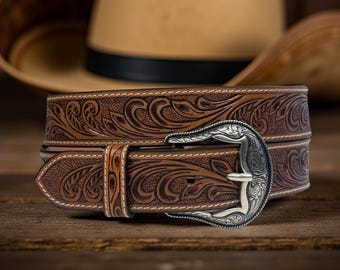 May include: Brown leather belt with intricate floral tooling and a silver-colored buckle. The belt features white stitching along the edges and a classic western design. A cowboy hat is in the background.