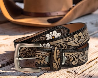 Op de afbeelding: Een bruine leren riem met een zilveren gesp en bloemenmotief. De riem heeft witte bloemaccenten en een gedetailleerd reliëfpatroon. Een bruine cowboyhoed staat op de achtergrond.