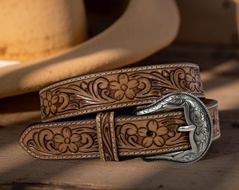 Op de afbeelding: Een lichtbruine leren riem met bloemmotieven en een zilverkleurige gesp. De riem heeft een gedetailleerd gesneden bloemmotief en een decoratieve gesp. De riem is waarschijnlijk 3,8 cm breed.