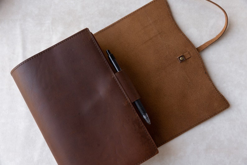 May include: A brown leather notebook with a pen holder and a strap closure. The notebook is open and the inside is lined with brown leather.