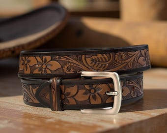 May include: A brown leather belt with floral and leaf tooling. The belt features a silver-colored buckle and is displayed on a wooden surface. The belt's design includes embossed flowers and foliage.
