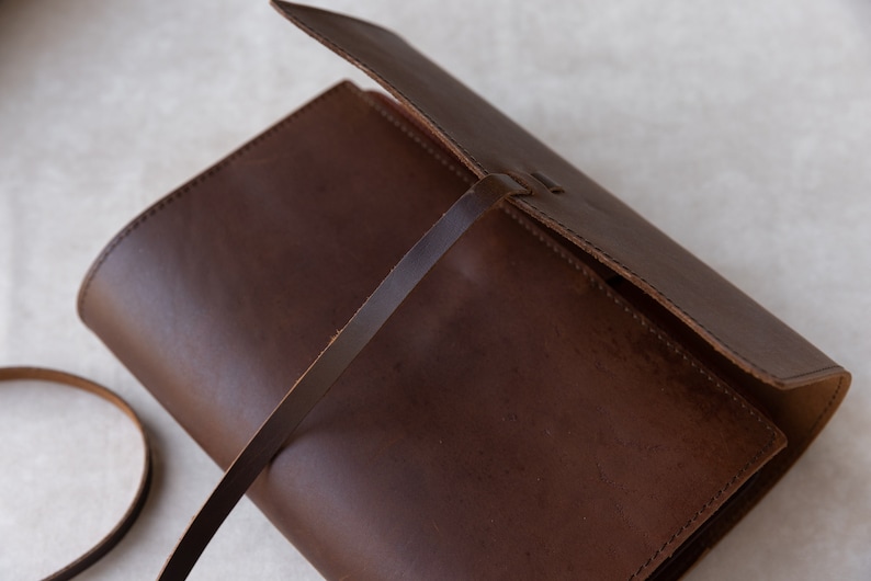 May include: A brown leather journal with a flap closure and a long brown leather strap. The journal is closed and the strap is wrapped around it.