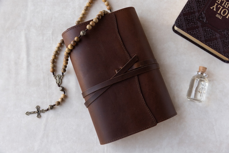 May include: A brown leather journal with a strap closure, a wooden rosary with a silver cross, a small glass bottle with a cork stopper, and a book titled "The Holy Bible".