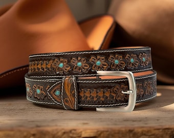 Op de afbeelding: Een bruine leren riem met bloemen en geometrische bewerkingen, turkooizen accenten en witte stiksels. De riem heeft een zilveren gesp. De riem is een modeaccessoire.