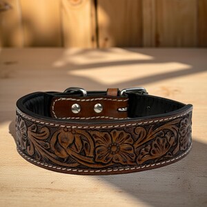 Op de afbeelding: Een bruine leren hondenhalsband met een bloemmotief. De halsband heeft een zwart interieur, zilverkleurige hardware en witte stiksels. De halsband wordt gepresenteerd op een houten oppervlak.