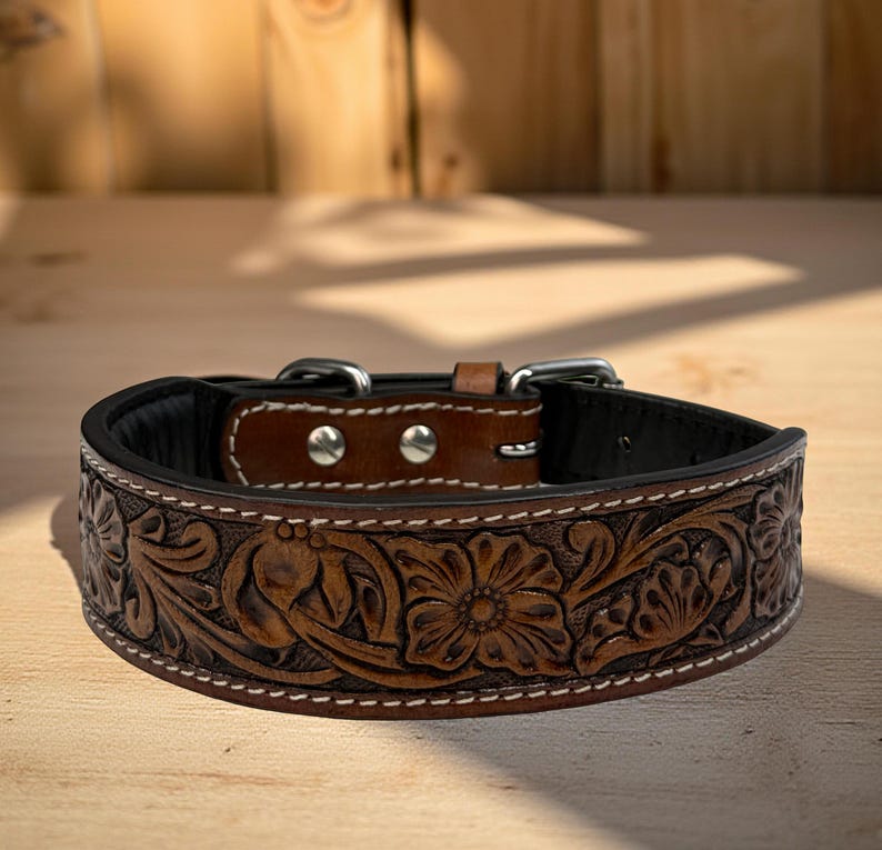 Op de afbeelding: Een bruine leren hondenhalsband met een bloemmotief. De halsband heeft een zwart interieur, zilverkleurige hardware en witte stiksels. De halsband wordt gepresenteerd op een houten oppervlak.