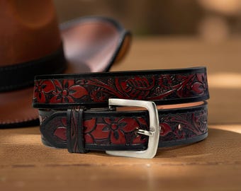 May include: A handcrafted leather belt with a floral design. The belt is black with a red floral pattern and a silver buckle. The belt is approximately 1.5 inches wide. A brown leather hat is in the background.