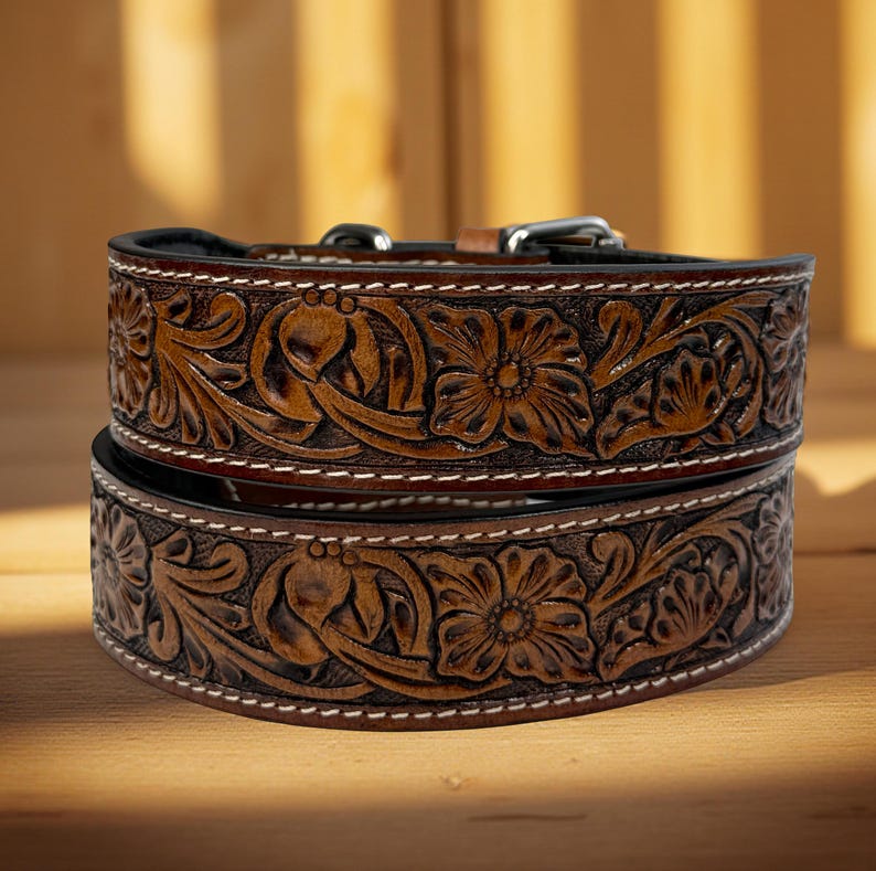 Op de afbeelding: Twee op elkaar gestapelde, bruine leren hondenhalsbanden met ingewikkelde bloemensnijwerken. De halsbanden hebben witte stiksels langs de randen en zilverkleurige metalen gespen. De gedetailleerde snijwerk en rijke bruine kleur geven de halsbanden een rustieke, handgemaakte uitstraling.