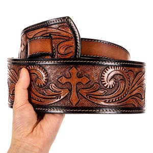 Pode incluir: Um cinto de couro castanho com uma cruz esculpida e um design floral. O cinto tem uma borda preta e é segurado por uma mão. É um acessório de estilo ocidental.
