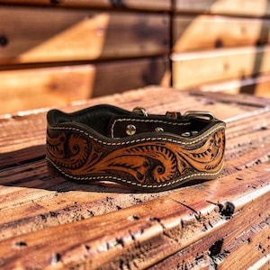 Peut inclure: Un collier de chien en cuir marron avec des gravures florales et tourbillonnantes complexes. Le collier pr&eacute;sente une bordure marron fonc&eacute; avec des coutures blanches et une boucle argent&eacute;e. Le collier est pos&eacute; sur une surface en bois.