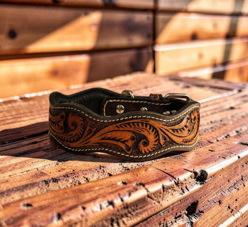 Peut inclure: Un collier de chien en cuir marron avec des gravures florales et tourbillonnantes complexes. Le collier pr&eacute;sente une bordure marron fonc&eacute; avec des coutures blanches et une boucle argent&eacute;e. Le collier est pos&eacute; sur une surface en bois.