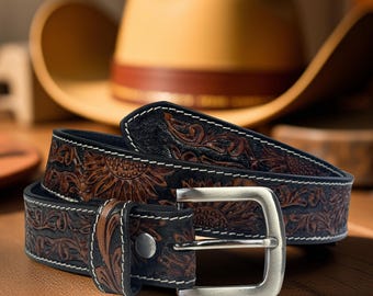 Puede incluir: Un cinturón de cuero marrón con un diseño de girasol y floral, costuras blancas y una hebilla plateada. El cinturón está sobre una superficie de madera, con un sombrero de vaquero en el fondo.