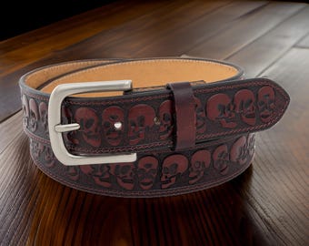 May include: A dark brown leather belt with a repeating skull pattern. The belt has a silver-colored buckle and a matching leather loop. The interior of the belt is a lighter tan color.
