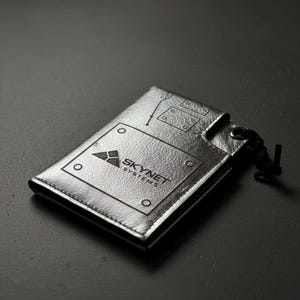Può includere: Portafoglio argentato con la scritta "SKYNET SYSTEMS" e un logo geometrico. Il portafoglio ha una superficie strutturata con dettagli simili a rivetti e un cordino nero. Design elegante e moderno.