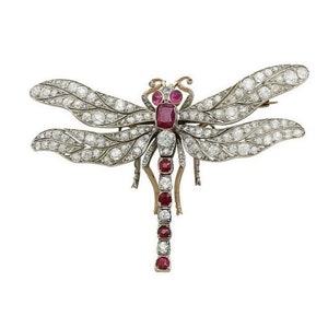 Puede incluir: Un broche de libélula de plata y rubí con un diseño detallado. Las alas están engastadas con diamantes y el cuerpo está adornado con rubíes.