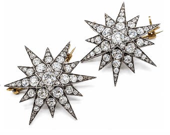 May include: Two star-shaped brooches featuring a multi-pointed design set with numerous clear, round-cut stones. The metal settings are dark, contrasting with the bright, reflective stones, and each brooch includes a gold-toned pin mechanism on the back.