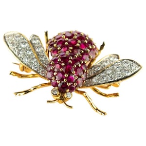 Puede incluir: Un broche de abeja de oro con detalles de rubí y diamantes. El cuerpo de la abeja está cubierto de rubíes y sus alas están engastadas con diamantes.