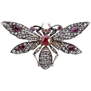 Puede incluir: Un broche de abeja de plata y oro con piedras preciosas rojas y blancas. Las alas de la abeja están extendidas y el cuerpo está cubierto de pequeñas piedras preciosas.